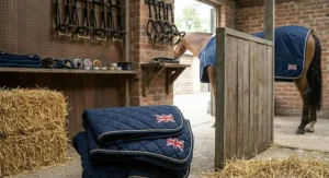A stack of folded navy quilted horse rugs embroidered with small Union Jack flags sitting on stable straw, with a rugged-up horse visible in the background stall.