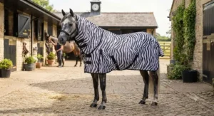 A grey pony standing in a stable yard wearing a zebra pattern combo fly rug for horses to deter flies naturally.